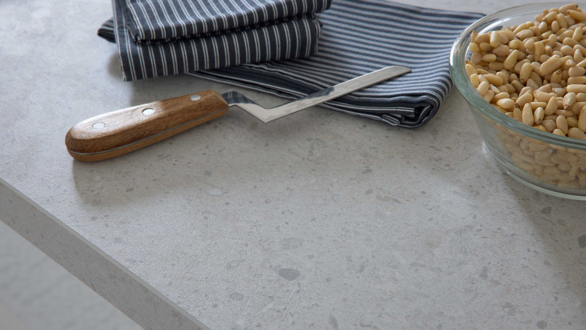 Auf einem hellen Tisch liegen gestreifte Küchentücher, ein Küchenmesser mit Holzgriff und eine Glasschüssel mit Pinienkernen.