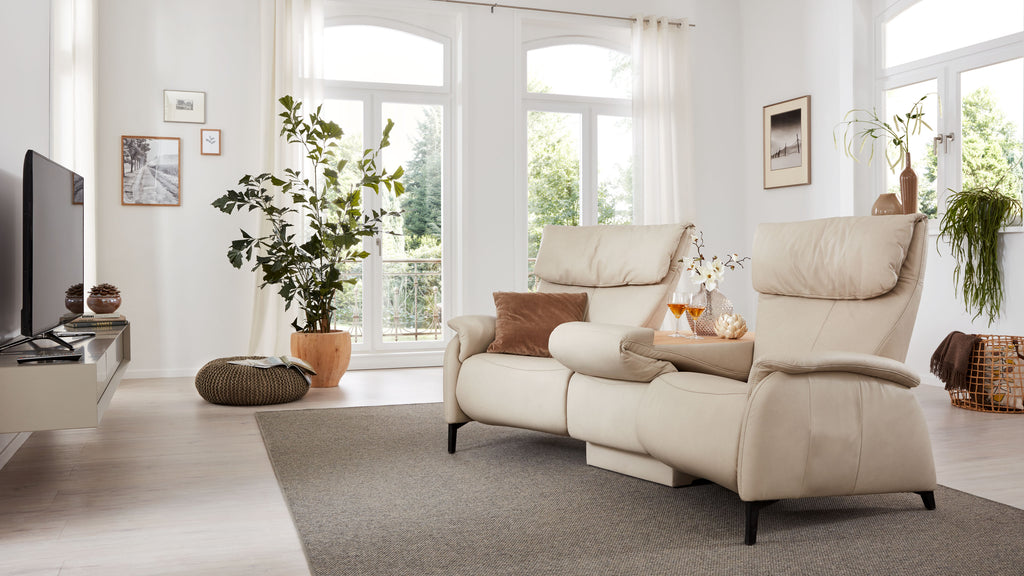 Beige Couch mit hohem Rücken und Kissen, vor Fenstern mit Blick ins Freie, umgeben von einer Pflanze und dekorativen Elementen.