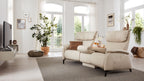Beige Couch mit hohem Rücken und Kissen, vor Fenstern mit Blick ins Freie, umgeben von einer Pflanze und dekorativen Elementen.