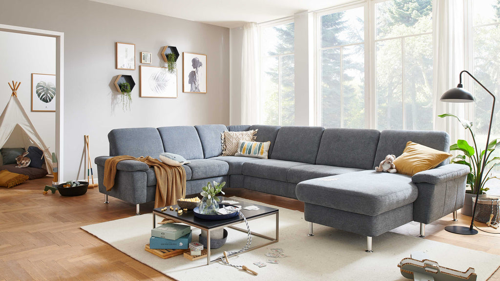 Eine graue Couch mit Kissen, ein Beistelltisch mit Pflanzen und Spielkarten sowie eine Stehlampe stehen in einem hellen Raum mit großen Fenstern.
