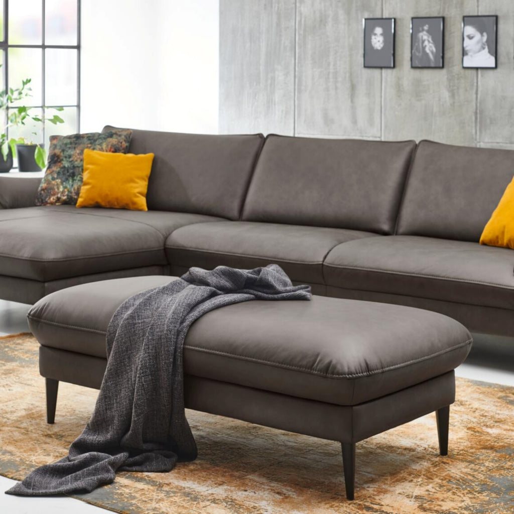 Modernes Wohnzimmer mit grauem Ecksofa, HUKLA® XXL-Hocker, gelben Kissen, einer Überwurfdecke und Wandbildern.