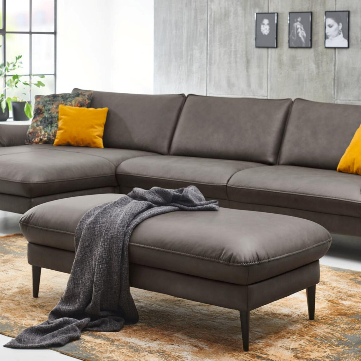 Modernes Wohnzimmer mit grauem Ecksofa, HUKLA® XXL-Hocker, gelben Kissen, einer Überwurfdecke und Wandbildern.