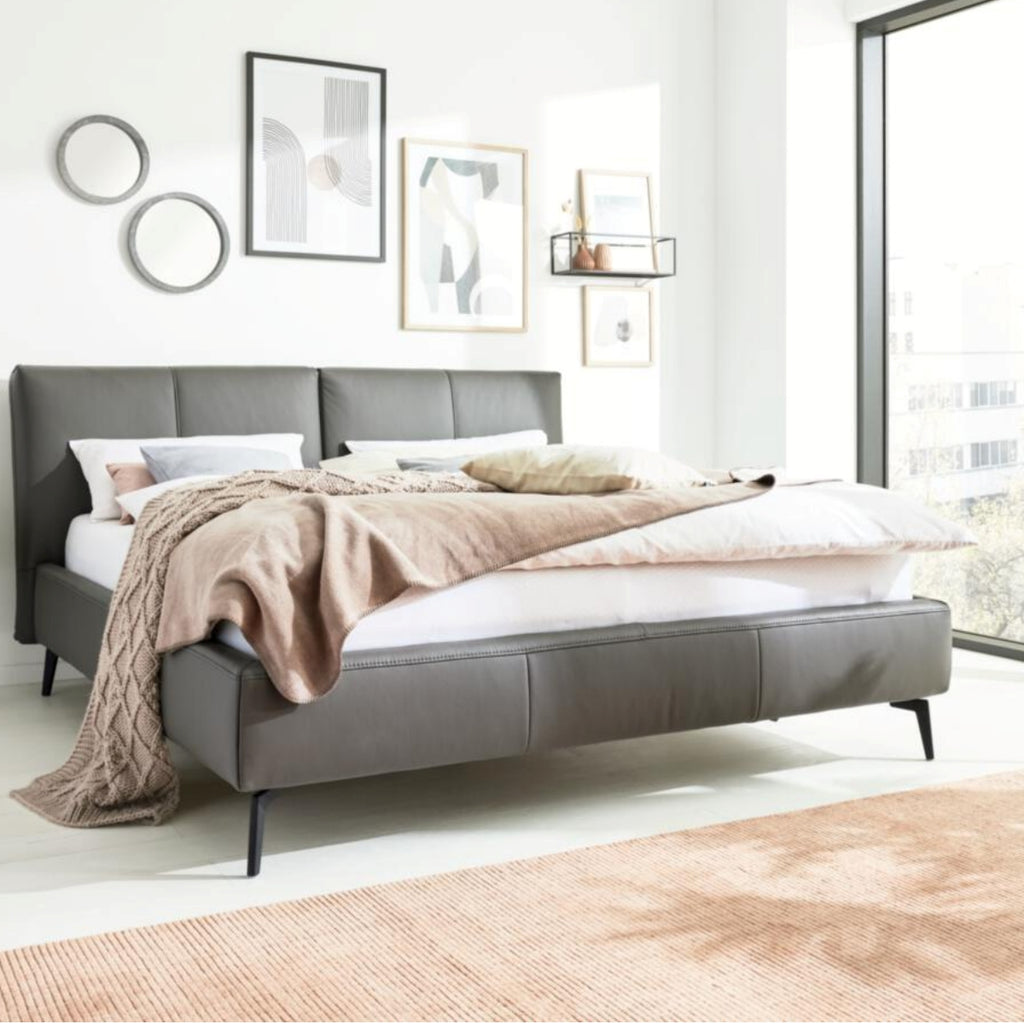 Modernes Schlafzimmer mit einem HUKLA® IL Interliving 1416 Lederbett 180 x 200 cm in Anthrazit, neutraler Bettwäsche, Kunstwerken und sonnendurchfluteten Fenstern.