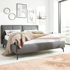 Modernes Schlafzimmer mit einem HUKLA® IL Interliving 1416 Lederbett 180 x 200 cm in Anthrazit, neutraler Bettwäsche, Kunstwerken und sonnendurchfluteten Fenstern.