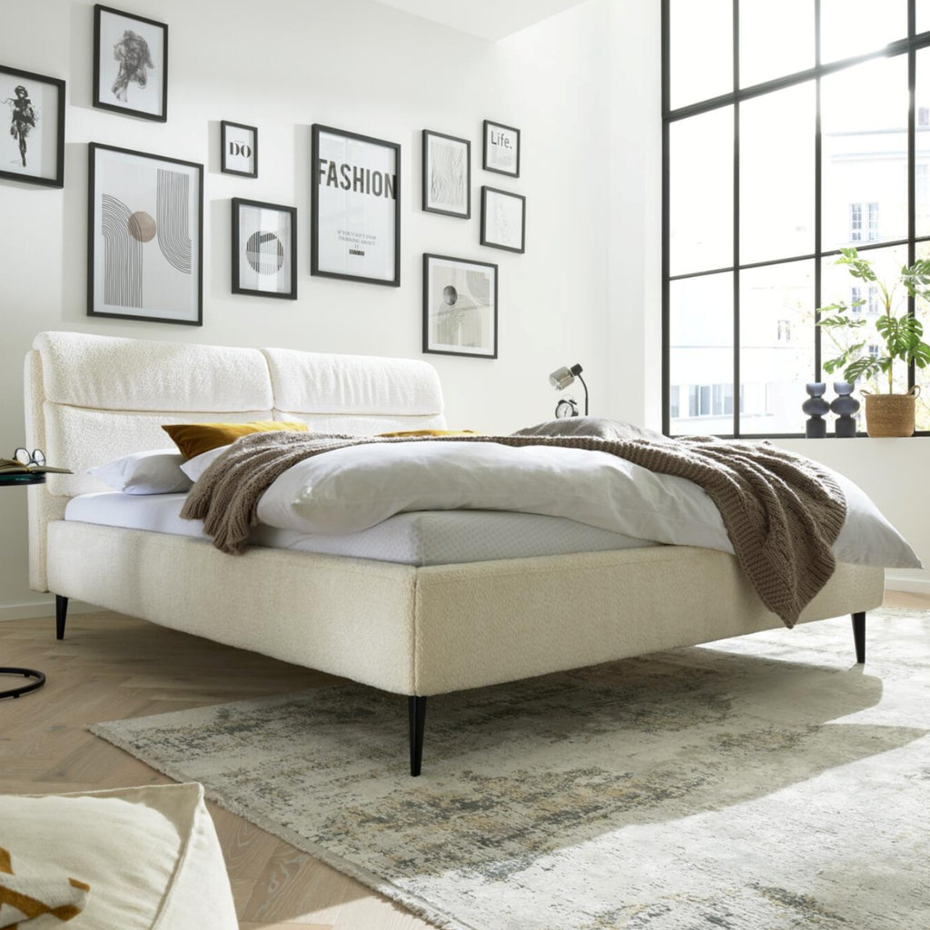 Modernes Schlafzimmer mit dem HUKLA® Interliving Serie 1416 Polsterbettgestell „Kama“ in Naturweiß, einer braunen Decke, Kunstwerken und Pflanzen.
