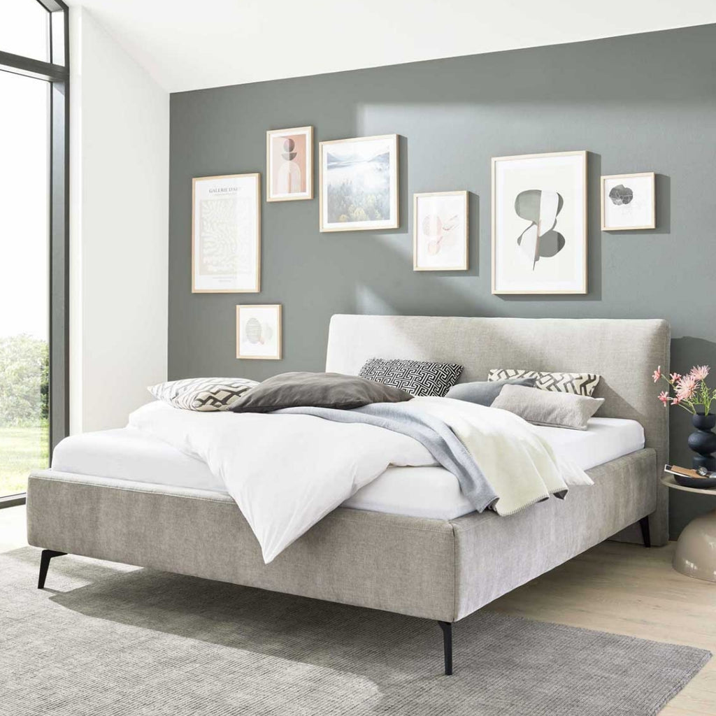 Ein modernes Schlafzimmer mit einem HUKLA®-Polsterbettgestell 180 × 200 cm, weißer Bettwäsche und Sonnenlicht, das durch große Fenster hereinströmt.