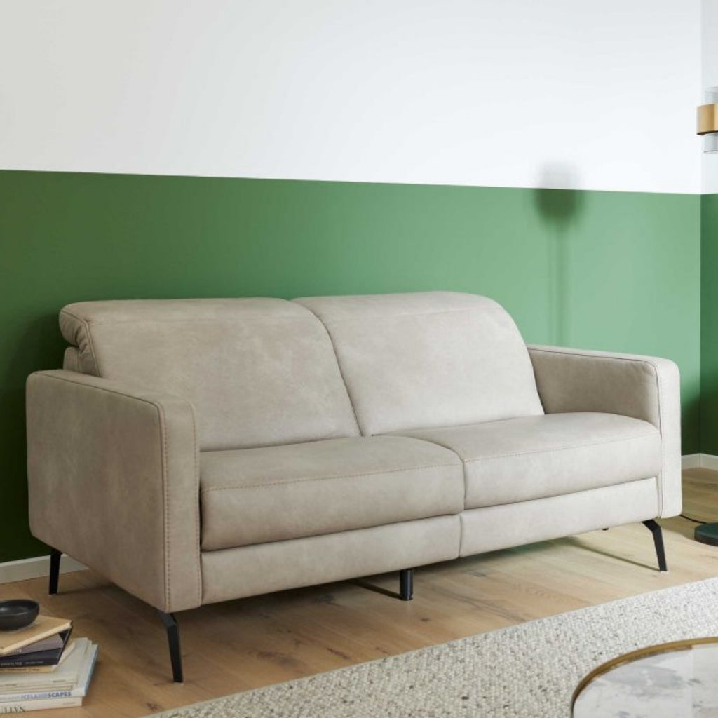 Beiges Sofa der Serie 4065 von Interliving, schwarze Metallbeine, daneben Bücher und ein runder Teppich auf hellem Holz vor einer grün-weißen Wand.