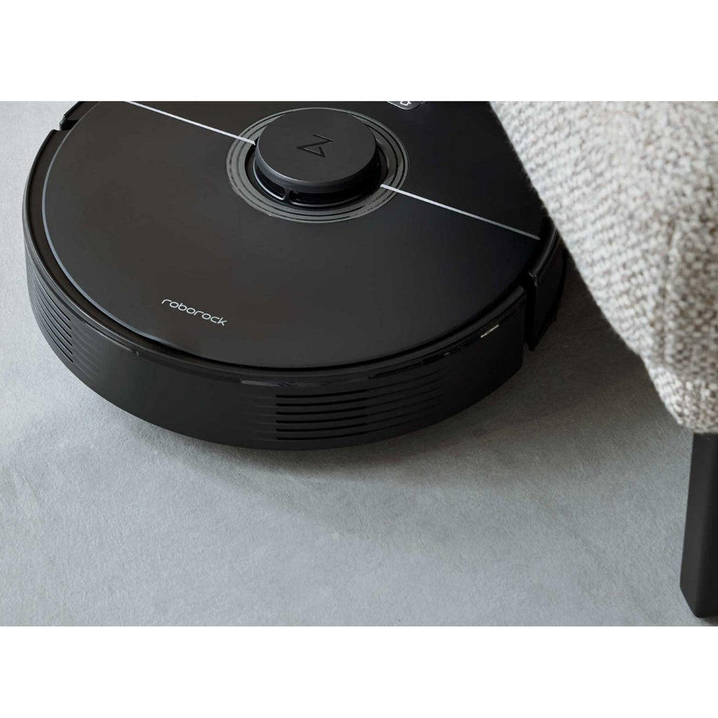 Ein schwarzer Staubsaugerroboter reinigt auf einem hellgrauen Boden neben dem Ecksofa „Interliving 4460“ in Silber von Steinpol Polsteria.