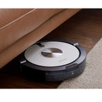 Ein ZACO-Staubsaugerroboter saugt unter einem Steinpol-Sofa in der Farbe „Haselnussbraun“ auf dem Holzboden neben einem beigen Teppich.