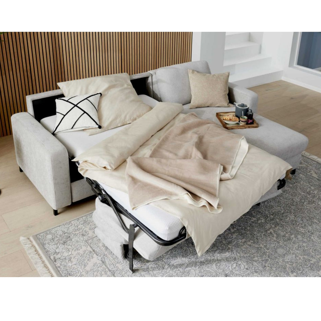HUKLA® IL Interliving 4700 Eckkombination in der Farbe „Toni Silver“, mit beiger Bettwäsche, Kissen, Tablett und Snacks auf einem hellen Teppich.