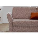Braunes Sofa der HUKLA® Interliving Serie 4700 mit einer Armlehne, orangefarbenem Kissen und hellem Holzboden im Hintergrund.