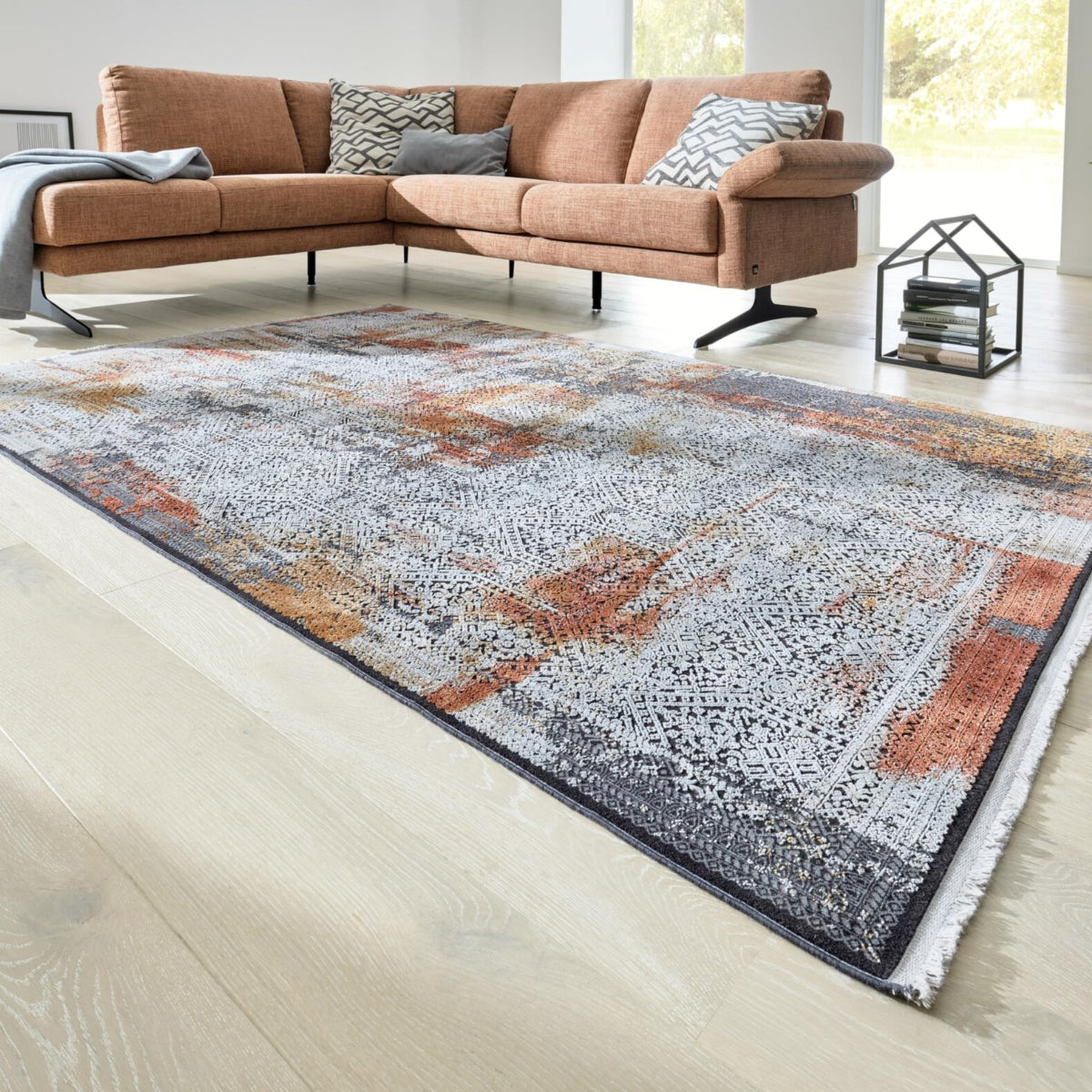 Ein Teppich „GOLZE | IL Interliving G-8060“ in Rost-, Sand- und Grautönen passt in diesem hellen Wohnzimmer perfekt zu einem orangefarbenen Sofa.
