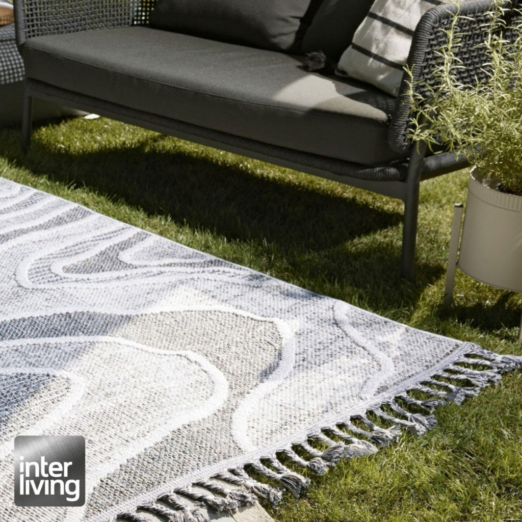 Outdoor-Teppich von GOLZE | IL in Hellgrau-Weiß mit modernem Muster und Fransen, auf grünem Rasen vor einem Sofa und einer Pflanze.