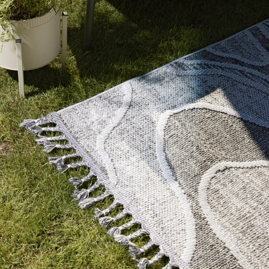 Der Outdoor-Teppich von GOLZE | IL in Hellgrau-Weiß liegt im Schatten auf dem Rasen neben einem Blumentopf und einem Tischbein.