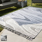 GOLZE | IL Outdoor-Teppich in Hellgrau-Weiß mit fließendem Muster und Fransen auf der Terrasse neben Möbeln und Rasen.