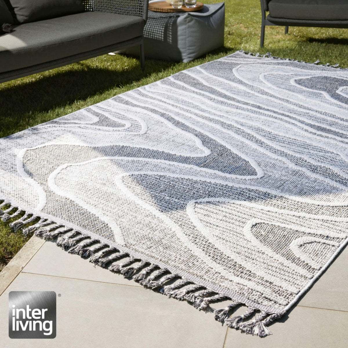 GOLZE | IL Outdoor-Teppich in Hellgrau-Weiß mit fließendem Muster und Fransen auf der Terrasse neben Möbeln und Rasen.