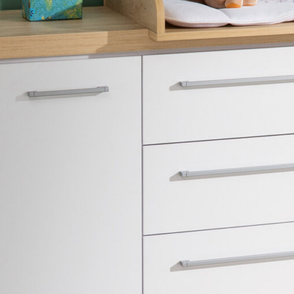 Weiße Kommode mit silbernen Griffen neben einem Kleiderschrank „Modell Kira“ von PAIDI 0190; in der Wickelkommode aus Holz befinden sich Taschentücher und eine Babymatte.