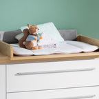 Stofftiere und Kissen auf einem gepolsterten Tisch mit weißen Schubladen, neben einem „0190 Kira“-Kleiderschrank im Kinderzimmer.
