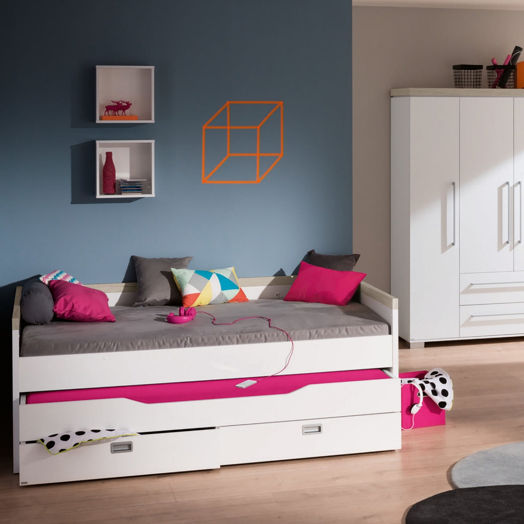 Modernes Schlafzimmer mit einem PAIDI Kira-Kojenbettgestell, rosa Bettwäsche, bunten Kissen und einem weißen Kleiderschrank vor einer blauen Wand.