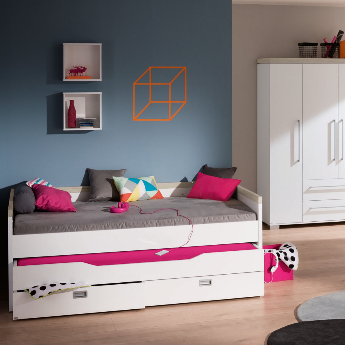 Modernes Schlafzimmer mit einem PAIDI Kira-Kojenbettgestell, rosa Bettwäsche, bunten Kissen und einem weißen Kleiderschrank vor einer blauen Wand.