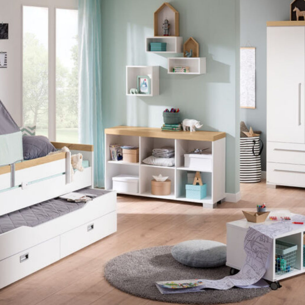 Modernes Kinderzimmer mit dem PAIDI Kira-Sideboard in Kreideweiß/Eiche, Regalen, einem weichen Teppich und Spielzeug auf einem Rolltisch.