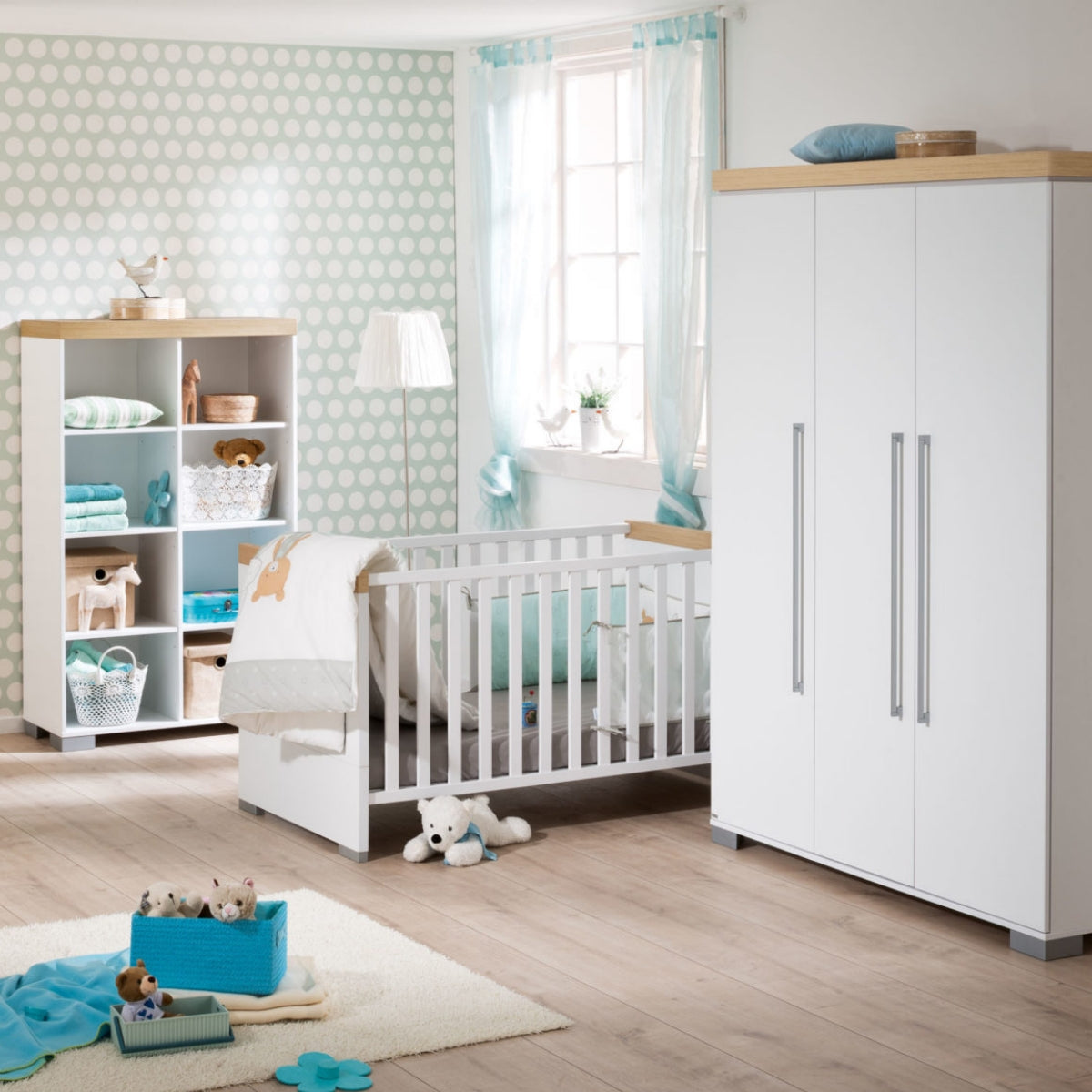 Modernes Kinderzimmer mit einem 0190 PAIDI Kira-Babybett, Spielzeug, blauen Akzenten, hellen Holzböden und Wänden mit Polka-Dot-Muster.
