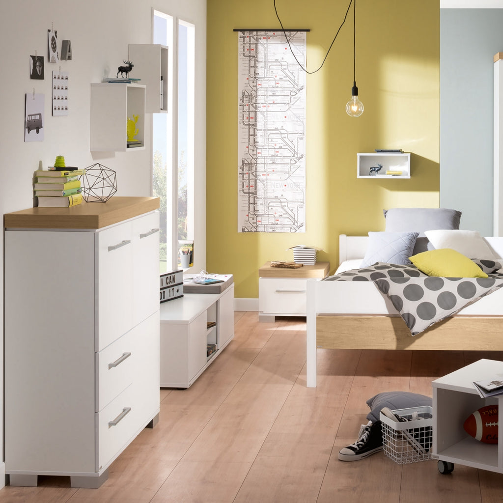 Modernes Schlafzimmer mit weißem PAIDI Kira-Hochschrank, gelber Akzentwand, Deko, Sportausrüstung und Holzboden.