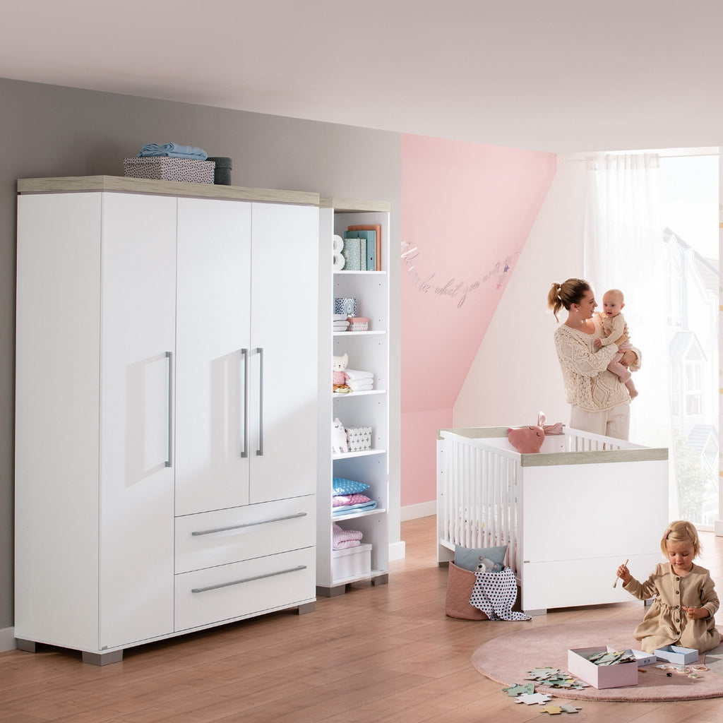 Eine Frau mit einem Baby steht neben einem Kinderbett in der Nähe eines PAIDI Kira Kleiderschranks, während ein Kind auf dem Boden spielt.