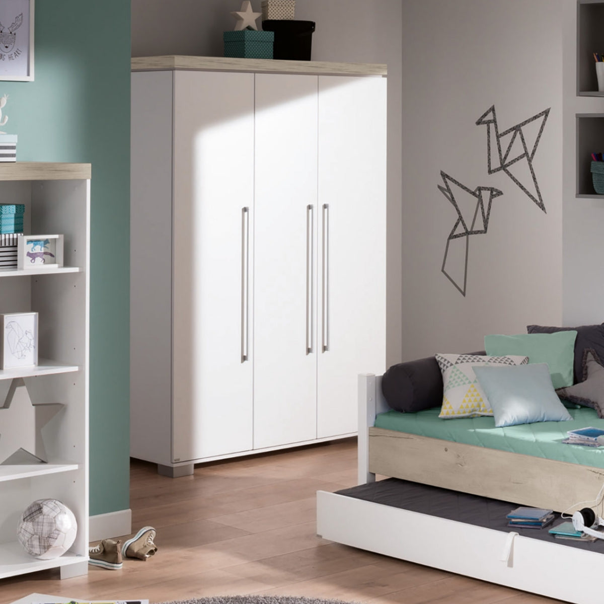 Das moderne Kinderzimmer mit türkisfarbenen Wänden ist mit einem Kleiderschrank „0190 PAIDI Kira 3T“, einem Tagesbett, Regalen und Origami-Vogel-Kunstwerken ausgestattet.