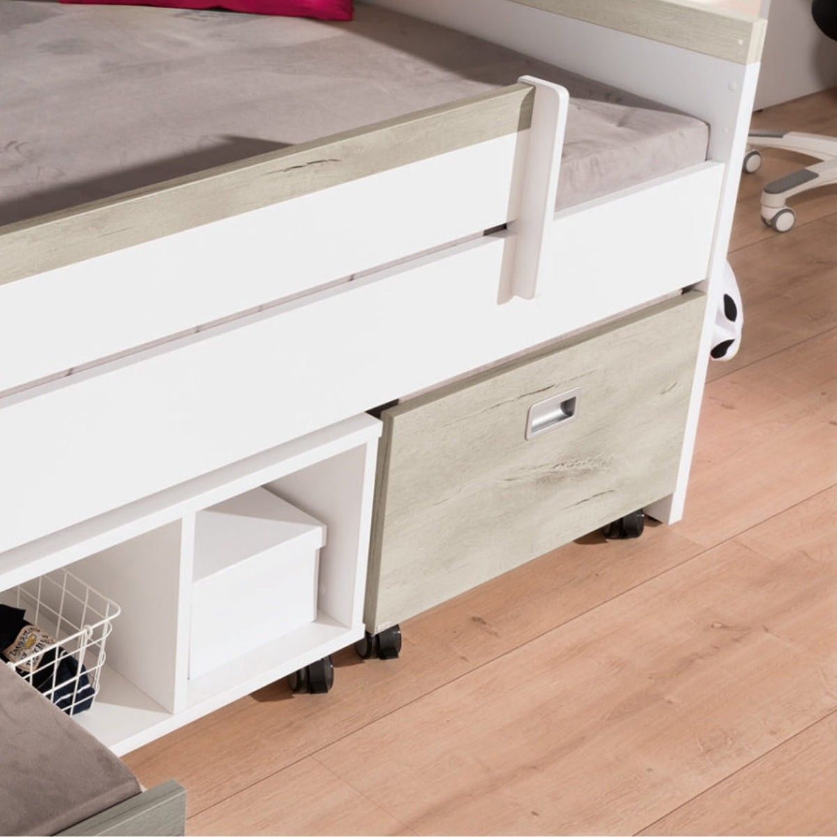 Ein Bett aus weißem Holz mit Stauraum, Fächern und einer PAIDI Kira Rollbox darunter, auf einem hellen Holzboden.