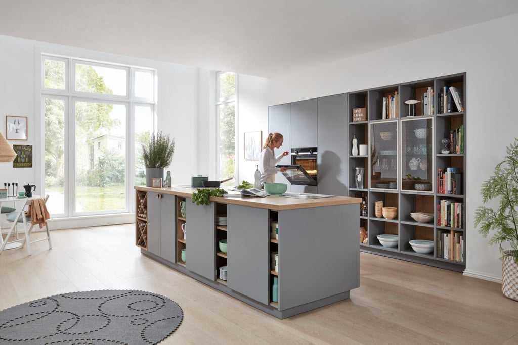 Eine Frau nutzt einen SIEMENS-Backofen in der WERT KÜCHE® Nora-Küche von schüller, die sich durch Grau- und Holzelemente sowie offene Regale auszeichnet.