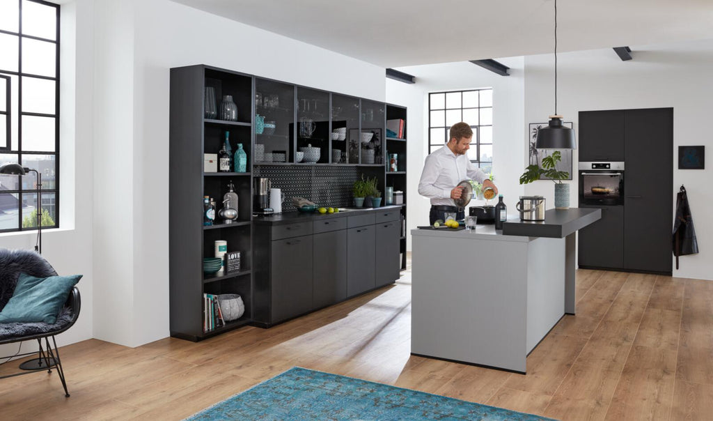 Ein Mann kocht in einer WERT KÜCHE® Franziska-Küche – elegant, schwarz, mit markantem Design und Sonnenlicht, das durch große Fenster hereinströmt.