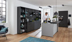 Ein Mann kocht in einer WERT KÜCHE® Franziska-Küche – elegant, schwarz, mit markantem Design und Sonnenlicht, das durch große Fenster hereinströmt.