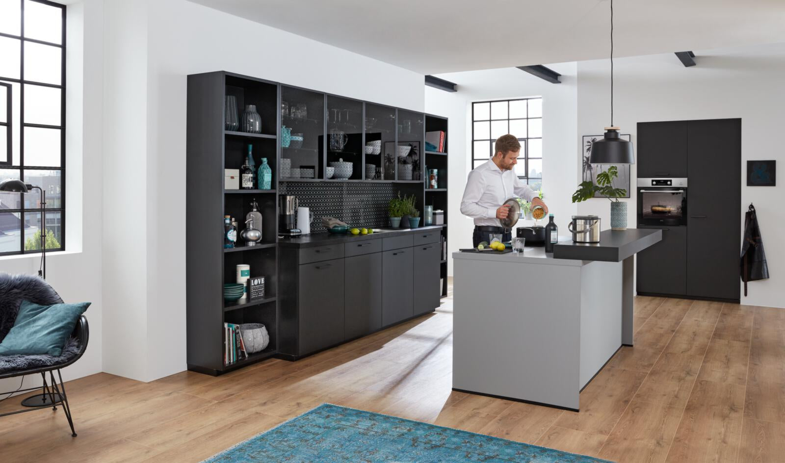 Ein Mann kocht in einer WERT KÜCHE® Franziska-Küche – elegant, schwarz, mit markantem Design und Sonnenlicht, das durch große Fenster hereinströmt.