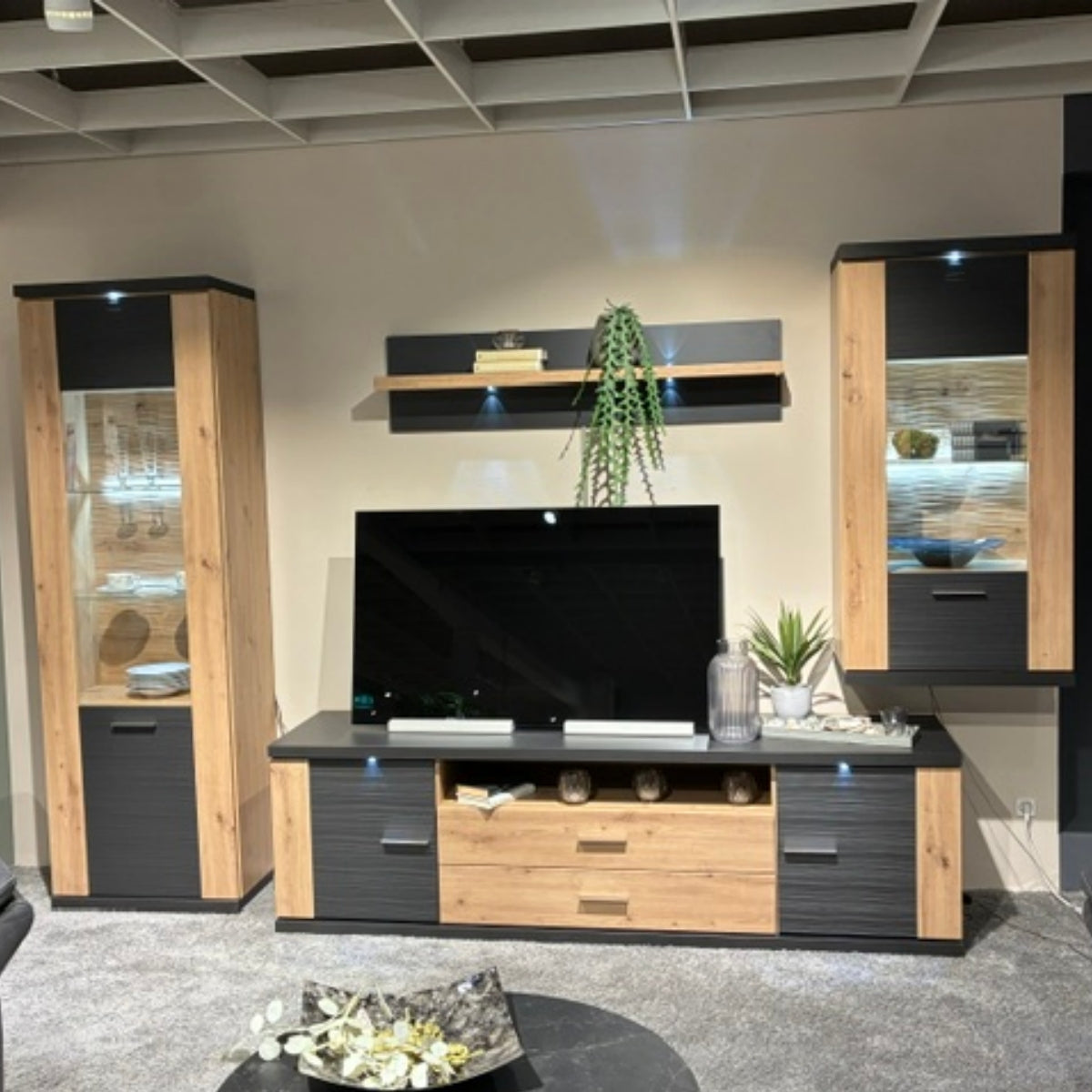 Modernes Wohnzimmer mit der Wohnkombination „Manhatten“ 0266, einem TV-Möbel, Regalen aus Eiche „Artisan“ und Zierpflanzen.