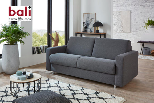 Bali Messina Funktionssofa – Schlafsofa von Bali in einem hellen Raum mit Pflanzen, Deko und dem „bali SITZEN LIEGEN“-Logo.