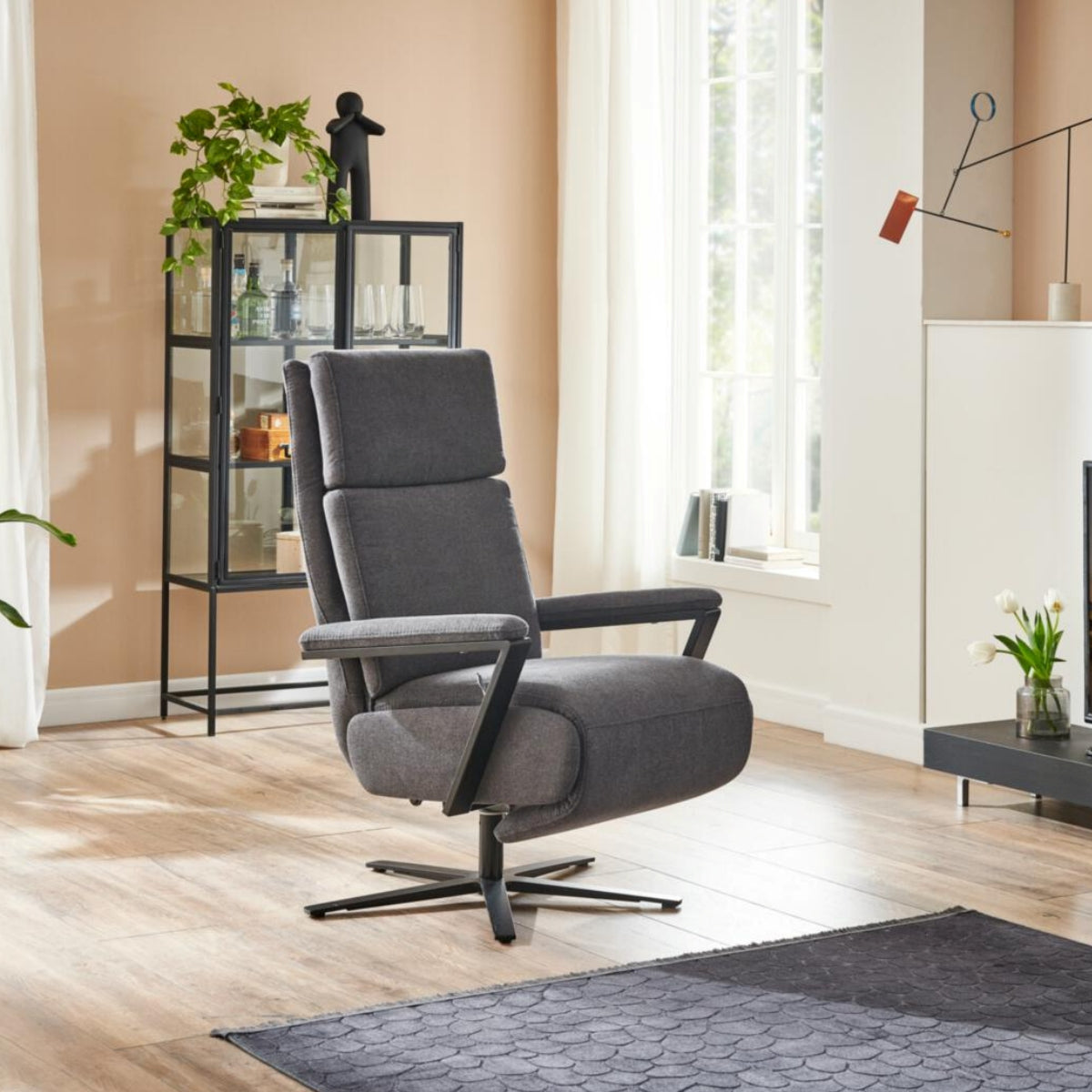 Ein grauer Relaxsessel L3 aus dem Polsterprogramm „OrthoSedis 4.0“ von Steinpol Polsteria steht auf einem Teppich in einem hellen Wohnzimmer.