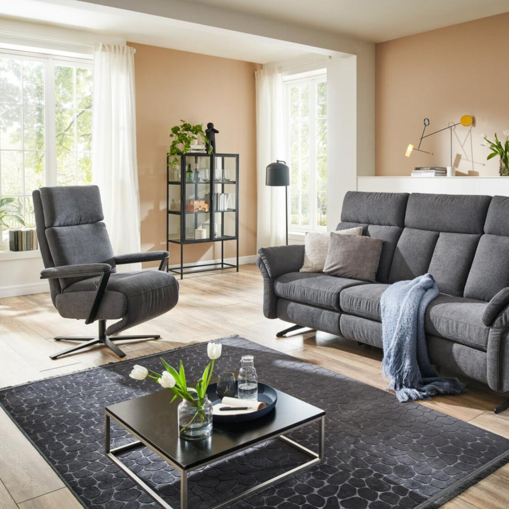 Modernes Wohnzimmer mit grauem Sofa, Relaxsessel „OrthoSedis 4.0 L3“ von Steinpol Polsteria, Couchtisch, Teppich, Pflanzen, Fenstern.