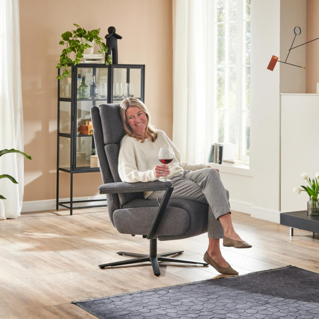 Eine Frau genießt ein Glas Rotwein und lächelt dabei auf einem „Steinpol Polsteria OrthoSedis 4.0 Relaxsessel L3“ in einem hellen Wohnzimmer.