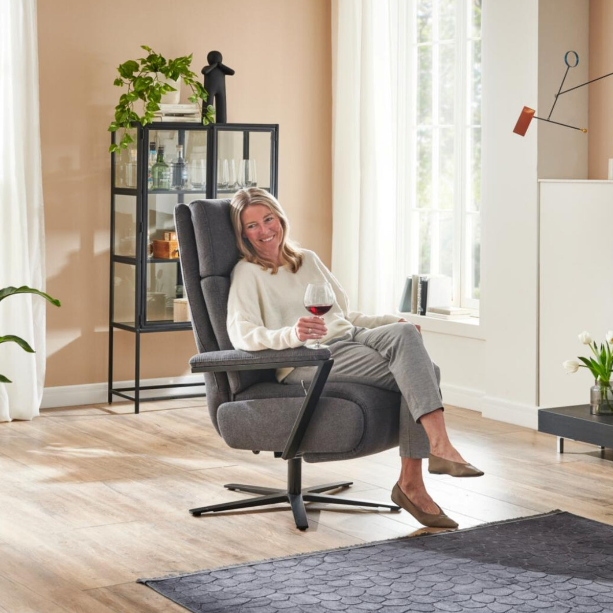 Eine Frau genießt ein Glas Rotwein und lächelt dabei auf einem „Steinpol Polsteria OrthoSedis 4.0 Relaxsessel L3“ in einem hellen Wohnzimmer.