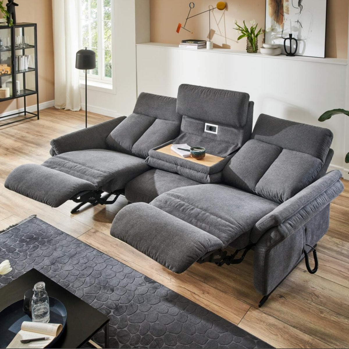 Zwei graue „OrthoSedis 4.0“-Rundsofas von Steinpol Polsteria mit Fußstützen und einer Konsole runden dieses sonnendurchflutete Wohnzimmer ab.