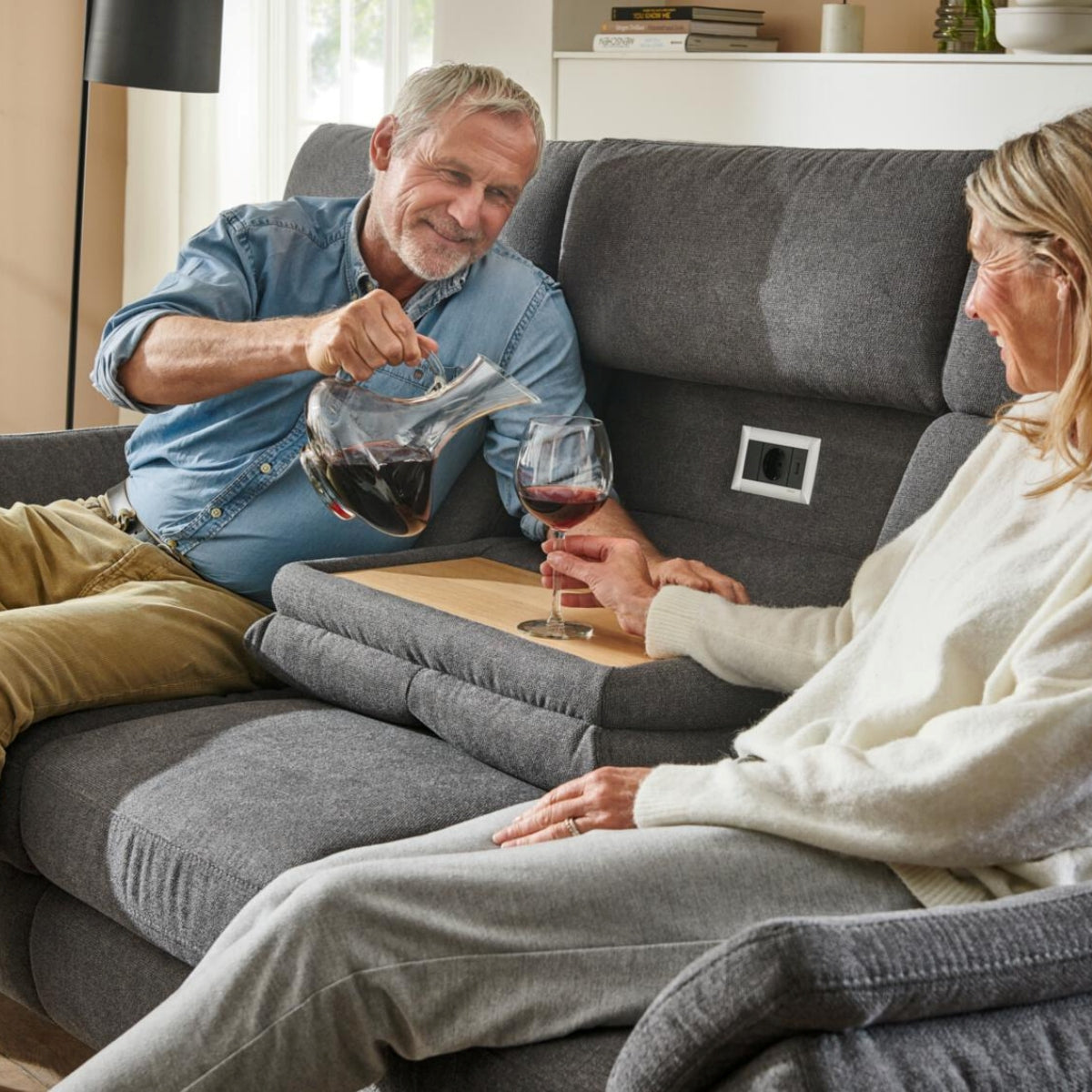 Ein Mann schenkt einer Frau Rotwein ein, während die beiden lächelnd auf einem grauen „Steinpol Polsteria OrthoSedis 4.0 Rundsofa“ sitzen.