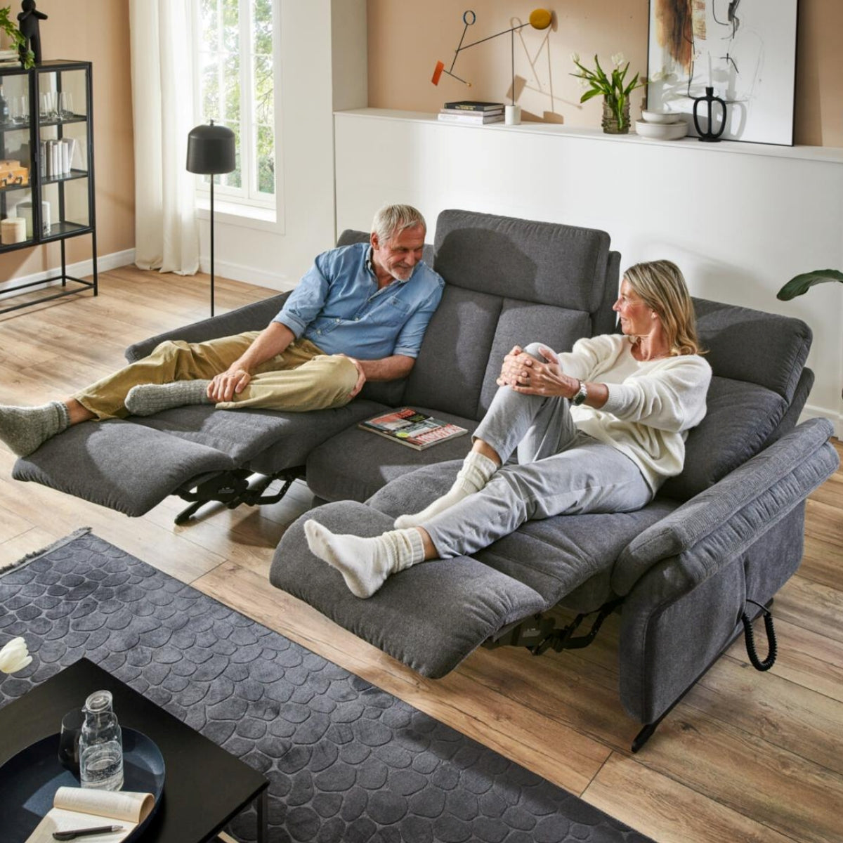 Zwei Personen entspannen sich auf dem Rundsofa „OrthoSedis 4.0“ von Steinpol Polsteria mit Federkern in einem hellen, gemütlichen Wohnzimmer.