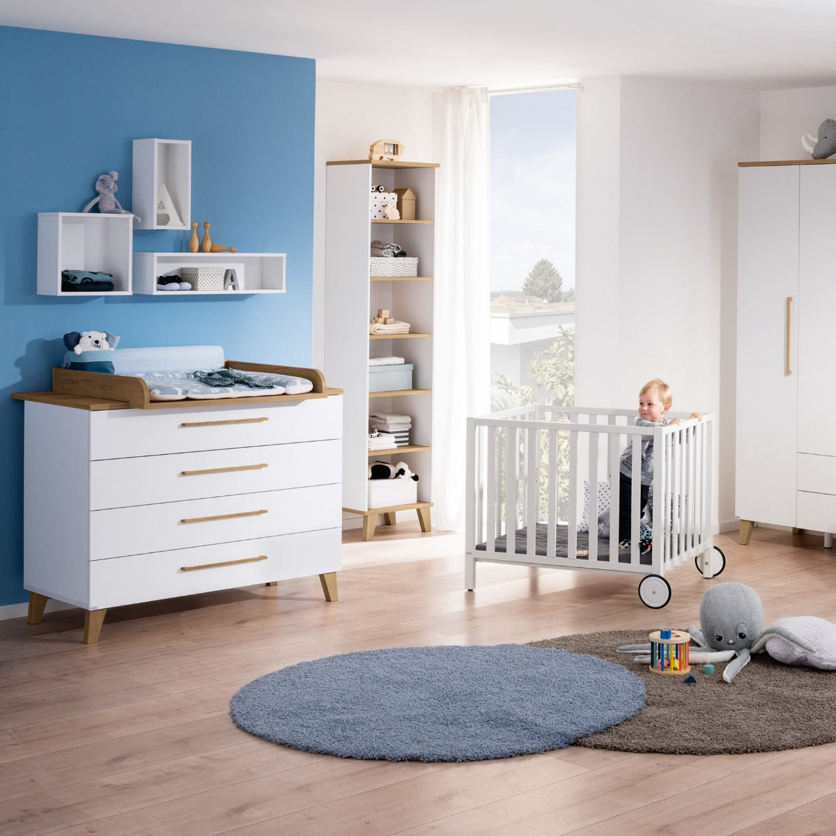 Ein Baby in einem weißen Kinderbett neben einer PAIDI Oscar-Schubladenkommode in Nebraska-Eiche-Optik mit blauen Akzenten.