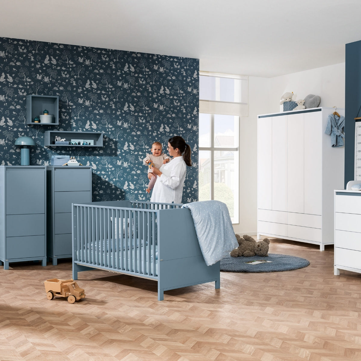 Eine Frau hält ein Baby im blauen PAIDI-Kinderzimmer mit der Oscar-Wandbox und dem Regal mit Stauraum von PAIDI im Arm.