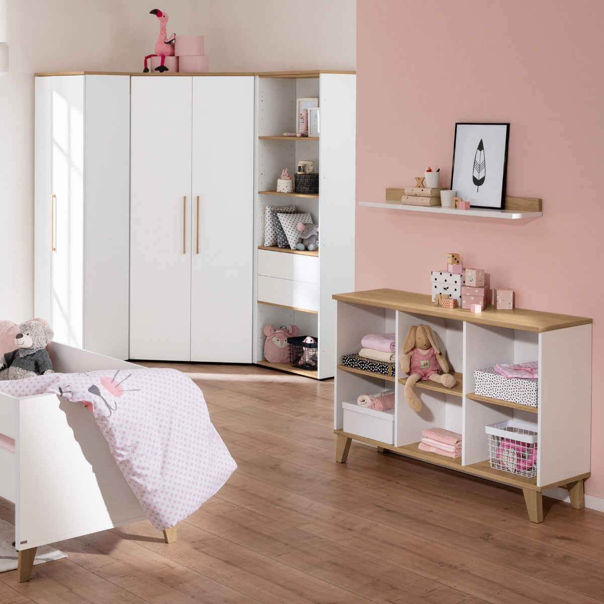 Modernes Kinderzimmer mit einem PAIDI Oscar Eck-Kleiderschrank in der Ausführung „Nebraska-Eiche“, rosa und weißer Einrichtung, Spielzeug und Regalen.