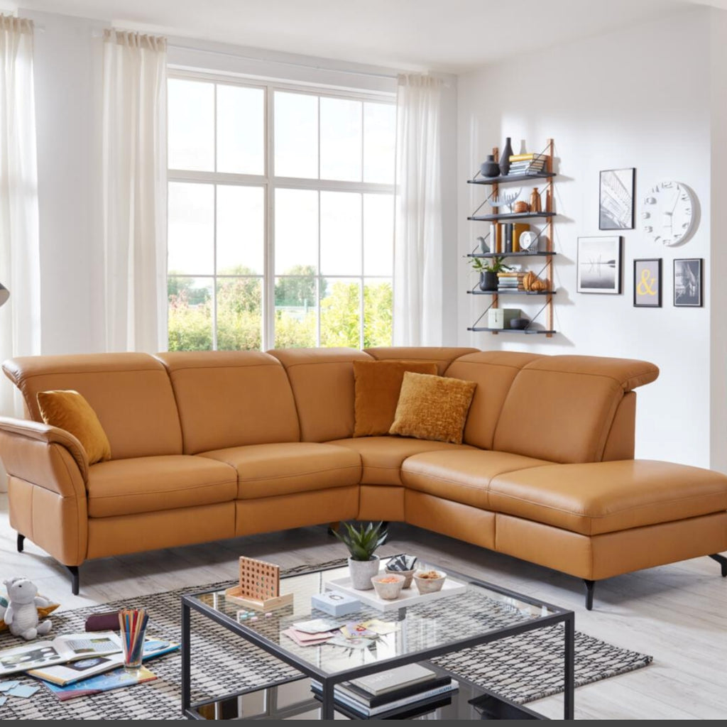 Wohnzimmer mit einem Ecksofa des Systems „Polsteria Senturo“ von Steinpol in senfgelbem Leder, großen Fenstern und stilvollen Wandregalen.