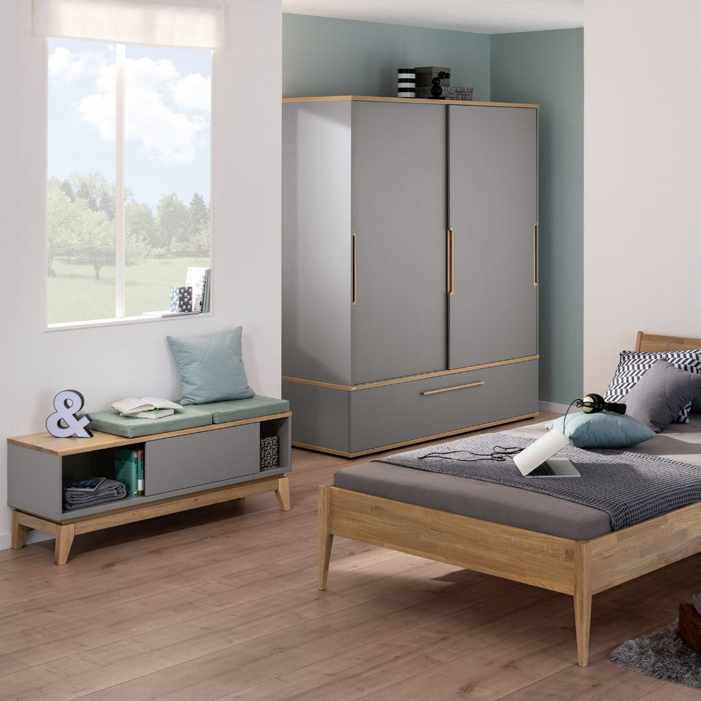 Modernes Schlafzimmer mit dem PAIDI Jugendzimmer-Lowboard „Sten“, schiefergrauen Melaminoberflächen, Holzboden und einer Fensterbank.