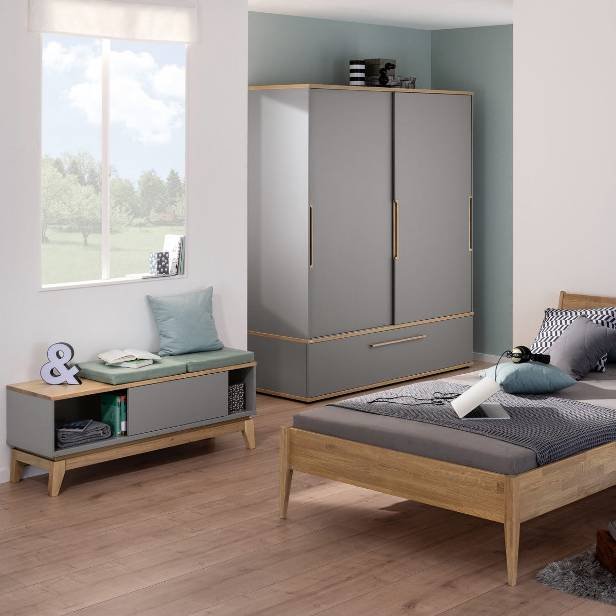 Modernes Schlafzimmer mit dem PAIDI Jugendzimmer-Lowboard „Sten“, schiefergrauen Melaminoberflächen, Holzboden und einer Fensterbank.