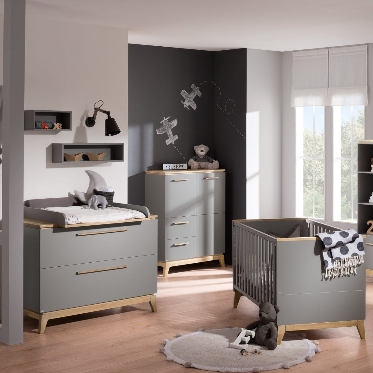 Kinderzimmer in Grau- und Holztönen mit der Wickelkommode „Sten“ von PAIDI, einem Kinderbett, Spielzeug, einer Wanddekoration mit Flugzeugmotiv und Eichenparkett.